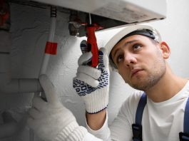 Plumber in a hard hat and gloves tightens a pipe under a sink with a wrench.