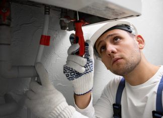 Instalación de calderas mejora eficiencia energética en hogares con asesoramiento técnico Plumber in a hard hat and gloves tightens a pipe under a sink with a wrench.