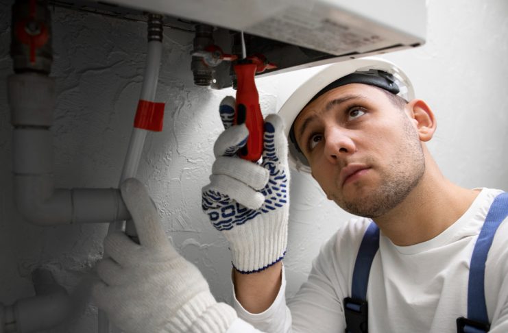 Plumber in a hard hat and gloves tightens a pipe under a sink with a wrench.