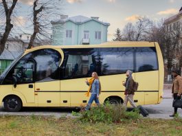 A yellow passenger bus on a city street with three pedestrians walking nearby, each carrying a drink or luggage as they pass.