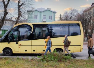 Alquiler de autocares crece como solución eficiente para la movilidad grupal A yellow passenger bus on a city street with three pedestrians walking nearby, each carrying a drink or luggage as they pass.