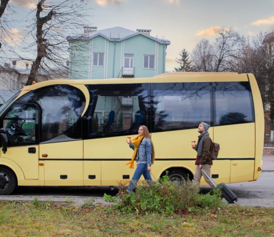 Alquiler de autocares crece como solución eficiente para la movilidad grupal A yellow passenger bus on a city street with three pedestrians walking nearby, each carrying a drink or luggage as they pass.