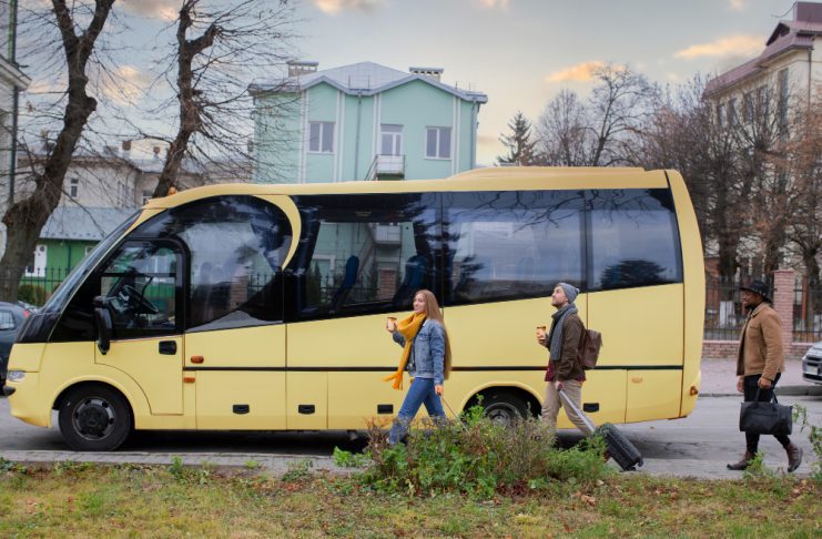 A yellow passenger bus on a city street with three pedestrians walking nearby, each carrying a drink or luggage as they pass.