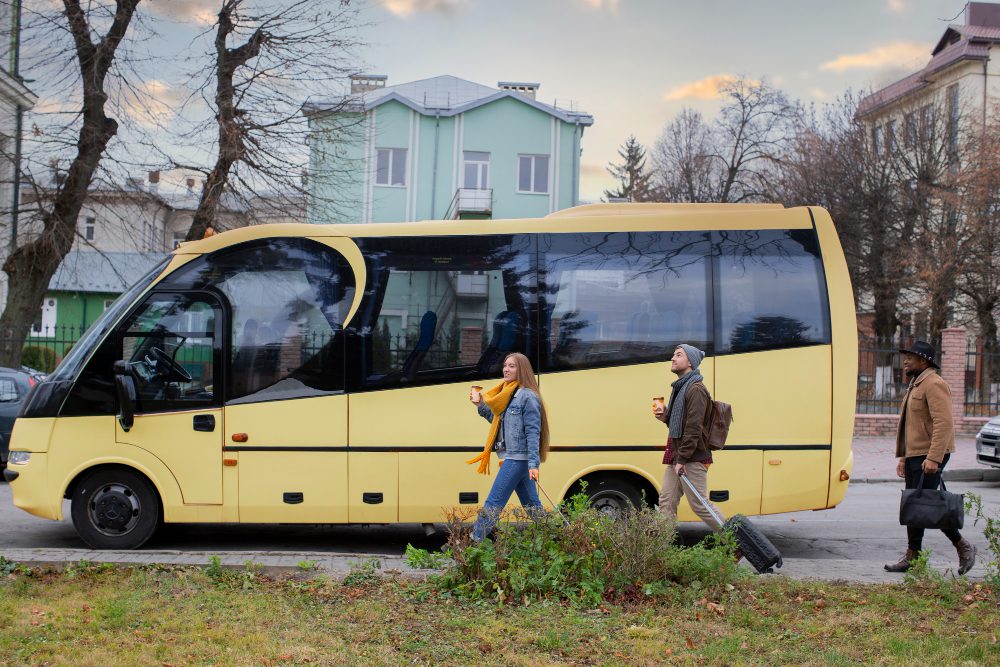 A yellow passenger bus on a city street with three pedestrians walking nearby, each carrying a drink or luggage as they pass.