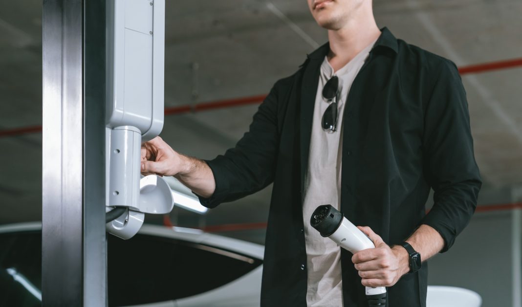 Person in a black shirt plugs in an electric vehicle charger at a car charging station in a parking garage, sunglasses tucked into shirt collar.