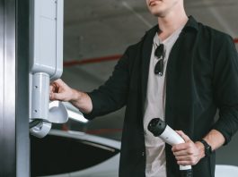 Person in a black shirt plugs in an electric vehicle charger at a car charging station in a parking garage, sunglasses tucked into shirt collar.
