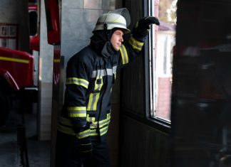 Soluciones de vidrio resistente al fuego mejoran seguridad en espacios profesionales Firefighter in full gear, helmet on, looking out a doorway toward a window inside a fire station with a red fire truck nearby