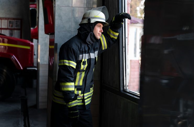Firefighter in full gear, helmet on, looking out a doorway toward a window inside a fire station with a red fire truck nearby