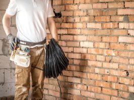 Construction worker in a white shirt and tool belt holding a coil of black cable against a brick wall.