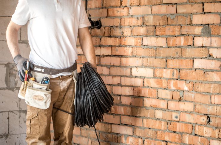 Construction worker in a white shirt and tool belt holding a coil of black cable against a brick wall.