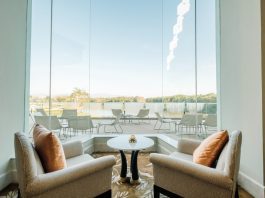 A pair of beige armchairs with orange cushions facing a small round table by a large glass window overlooking an outdoor patio and lake.