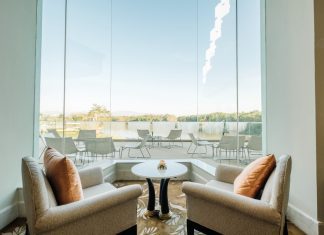 Los ventanales de gran formato marcan la evolución del diseño residencial A pair of beige armchairs with orange cushions facing a small round table by a large glass window overlooking an outdoor patio and lake.