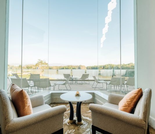 Los ventanales de gran formato marcan la evolución del diseño residencial A pair of beige armchairs with orange cushions facing a small round table by a large glass window overlooking an outdoor patio and lake.