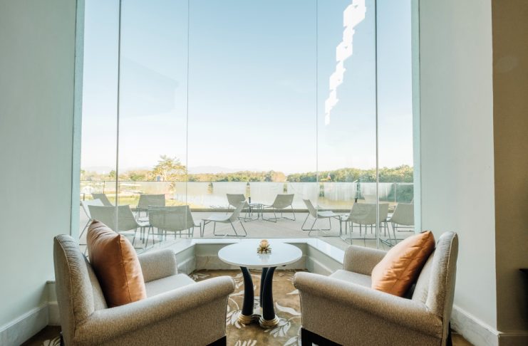 A pair of beige armchairs with orange cushions facing a small round table by a large glass window overlooking an outdoor patio and lake.