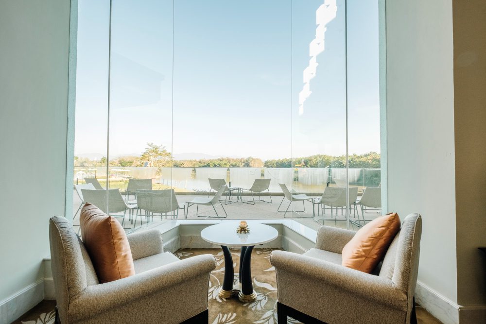 Los ventanales de gran formato marcan la evolución del diseño residencial A pair of beige armchairs with orange cushions facing a small round table by a large glass window overlooking an outdoor patio and lake.