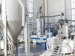 Industrial processing plant interior with tall stainless steel tanks, connected pipes, and control panels.