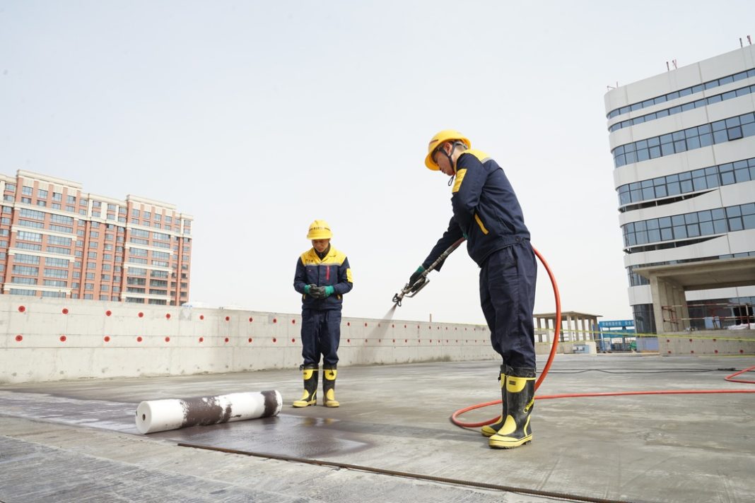 Two construction workers in yellow hard hats and dark blue safety outfits on a rooftop; one uses a spraying tool connected to a red hose, while a rolled membrane lies on the ground nearby.