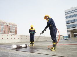 Two construction workers in yellow hard hats and dark blue safety outfits on a rooftop; one uses a spraying tool connected to a red hose, while a rolled membrane lies on the ground nearby.