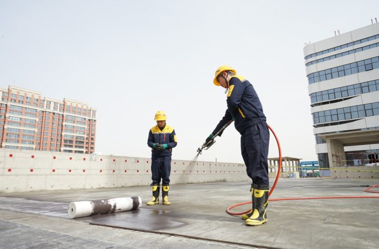 Two construction workers in yellow hard hats and dark blue safety outfits on a rooftop; one uses a spraying tool connected to a red hose, while a rolled membrane lies on the ground nearby.