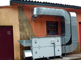 Outdoor industrial HVAC system: large galvanized ducts connected to a white air handling unit against an orange wall with a barred window and stone column nearby.