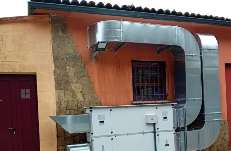 Outdoor industrial HVAC system: large galvanized ducts connected to a white air handling unit against an orange wall with a barred window and stone column nearby.