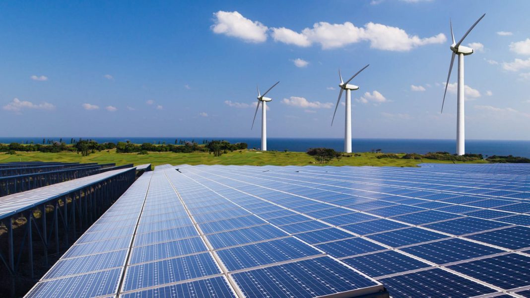 Los avances en la ingeniería impulsan la eficiencia en el transporte y la generación de energía limpia Rows of solar panels extend toward the horizon with three wind turbines on a grassy ridge and the sea in the background.