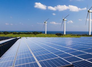 Los avances en la ingeniería impulsan la eficiencia en el transporte y la generación de energía limpia Rows of solar panels extend toward the horizon with three wind turbines on a grassy ridge and the sea in the background.