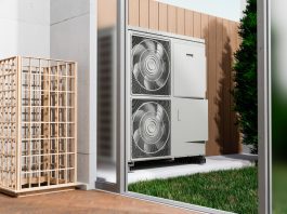 Empresas especializadas acercan soluciones de climatización para proyectos cada vez más eficientes - Outdoor HVAC condenser unit with two large fans mounted inside a glass enclosure beside a wooden privacy screen, with grass nearby.