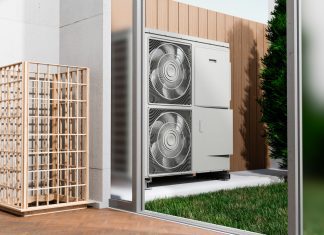 Empresas especializadas acercan soluciones de climatización para proyectos cada vez más eficientes Empresas especializadas acercan soluciones de climatización para proyectos cada vez más eficientes - Outdoor HVAC condenser unit with two large fans mounted inside a glass enclosure beside a wooden privacy screen, with grass nearby.