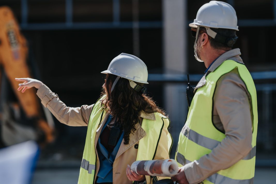 Two construction workers in hard hats and high-visibility vests discuss plans on a building site; the woman points while the man holds rolled blueprints at her side.