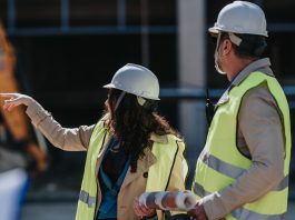 Two construction workers in hard hats and high-visibility vests discuss plans on a building site; the woman points while the man holds rolled blueprints at her side.