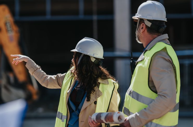 Two construction workers in hard hats and high-visibility vests discuss plans on a building site; the woman points while the man holds rolled blueprints at her side.