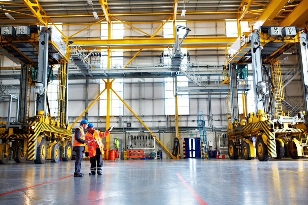 Tres proyectos clave de la ingeniería industrial Two engineers in safety vests discuss plans inside a large industrial facility with yellow overhead cranes and machinery nearby.