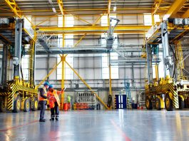 Two engineers in safety vests discuss plans inside a large industrial facility with yellow overhead cranes and machinery nearby.