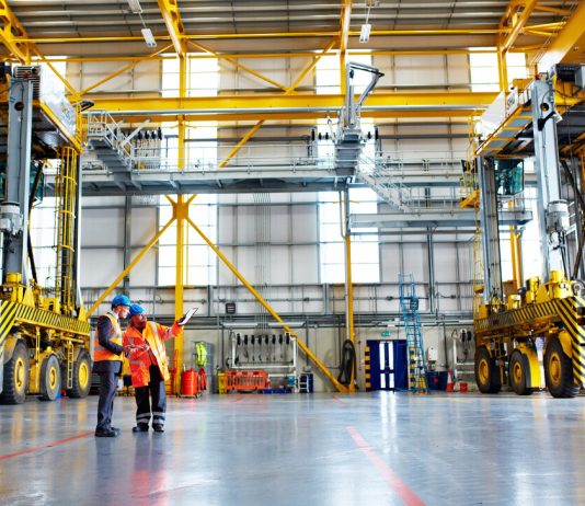 Tres proyectos clave de la ingeniería industrial Two engineers in safety vests discuss plans inside a large industrial facility with yellow overhead cranes and machinery nearby.