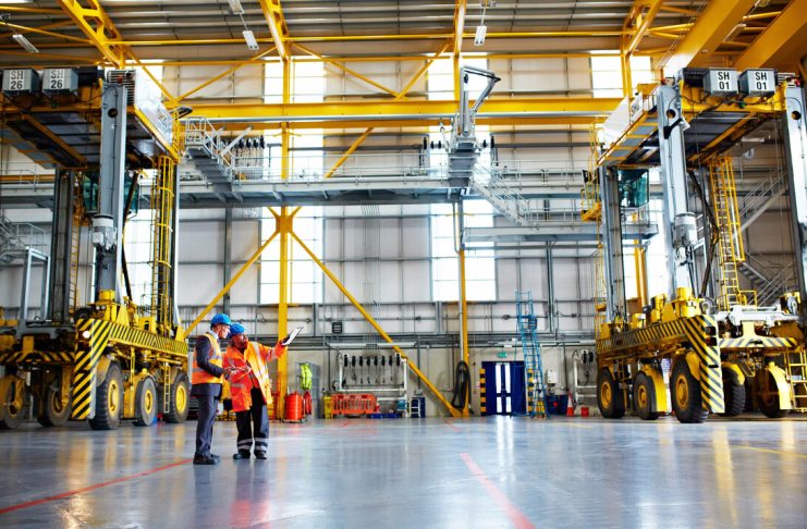 Two engineers in safety vests discuss plans inside a large industrial facility with yellow overhead cranes and machinery nearby.